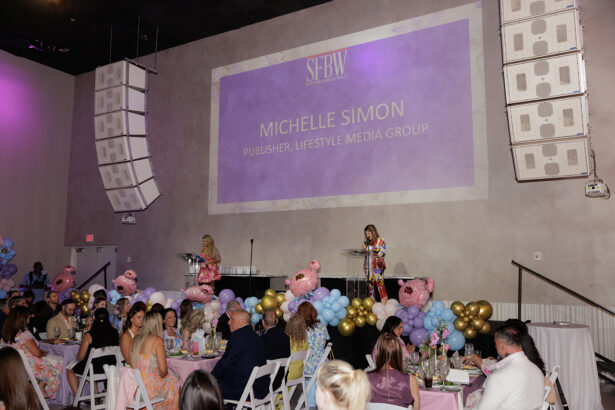 A large indoor event with people seated at decorated tables, watching two women speaking on stage. The screen behind them displays “Michelle Simon, Publisher, Lifestyle Media Group” at an SFBW event. South Florida Business & Wealth