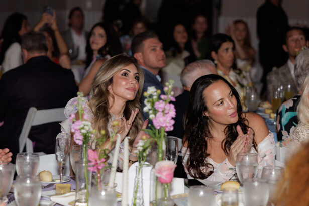 A group of people, seated at tables decorated with flowers and glasses, attentively watch and applaud during a formal indoor event. Two women in the foreground appear focused and are clapping. South Florida Business & Wealth
