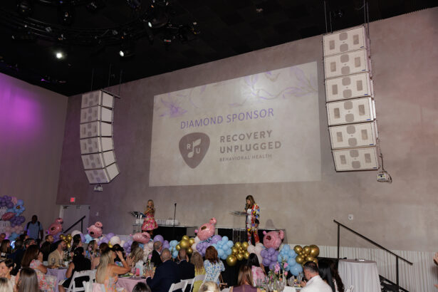 A large indoor event with people seated at decorated tables. Two women stand on stage with microphones. A projected sign reads "Diamond Sponsor Recovery Unplugged Behavioral Health." Balloons and teddy bears decorate the area. South Florida Business & Wealth