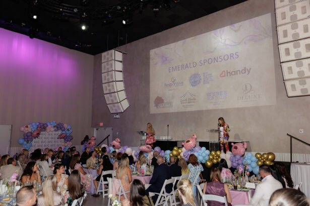 A large indoor event with seated guests watching speakers on stage. The wall displays "Emerald Sponsors" and sponsor logos. Tables have pastel decorations, and there are balloon arrangements and pink bear figures. South Florida Business & Wealth