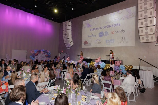 A large group of people sit at round tables with floral centerpieces at a formal indoor event, listening to two women speaking on a decorated stage. A screen behind them displays “Emerald Sponsors” and various sponsor logos. South Florida Business & Wealth