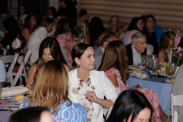 A group of people sit at round tables covered with blue tablecloths, eating and conversing at an indoor event. A woman in a white blazer with a name tag is in focus, looking to her left and smiling. South Florida Business & Wealth