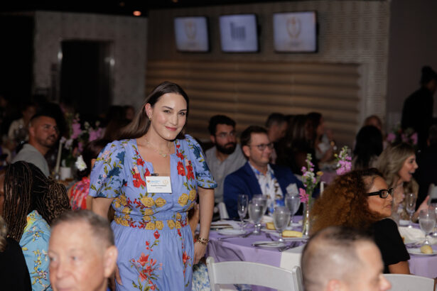 A woman in a blue floral dress stands and smiles among seated guests at a formal event with decorated tables and flower centerpieces. Several people are looking toward her. South Florida Business & Wealth