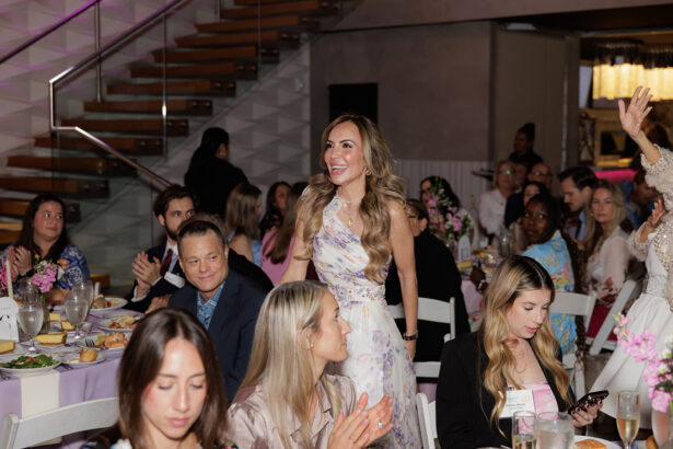 A woman in a floral dress stands and smiles as she’s applauded by people seated around tables at a formal indoor event. The room is decorated with flowers, and a staircase is visible in the background. South Florida Business & Wealth