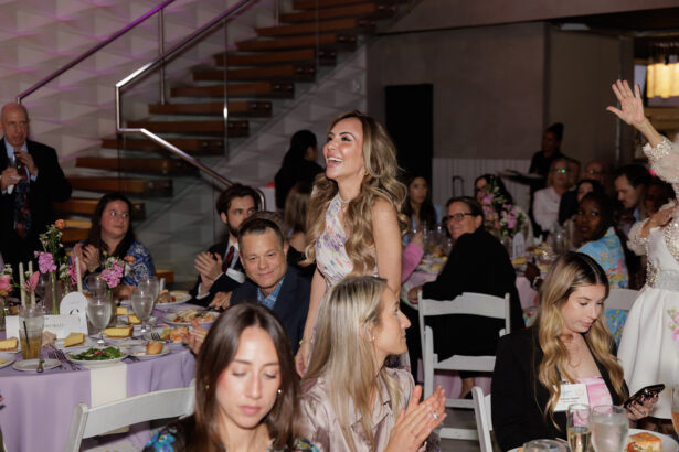 A woman stands smiling as others seated around banquet tables with lavender tablecloths applaud her at an indoor event; the atmosphere appears celebratory and formal. South Florida Business & Wealth