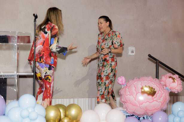 Two women in colorful floral outfits greet each other enthusiastically on a stage decorated with pastel balloons and large pink flower props, next to a clear podium and microphone. South Florida Business & Wealth