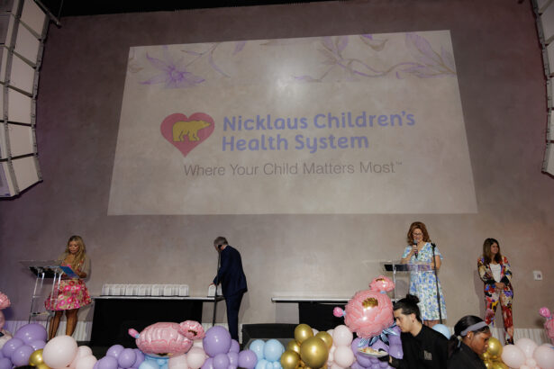 Four people stand on a stage decorated with pastel balloons and animal figures. Behind them, a projection displays the Nicklaus Children’s Health System logo and slogan: "Where Your Child Matters Most. South Florida Business & Wealth