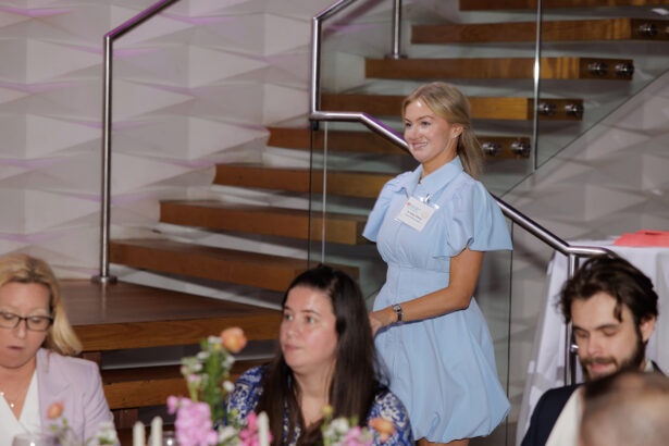 A woman in a light blue dress stands and smiles near a staircase at an indoor event, while other seated guests talk at a table decorated with flowers. South Florida Business & Wealth