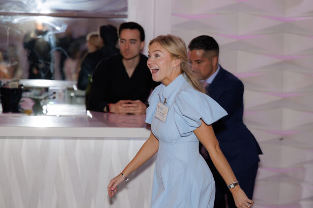 A smiling woman in a light blue dress walks indoors, looking excited. Two men stand in the background, one leaning on a white counter and another in a suit, with a geometric white wall behind them. South Florida Business & Wealth