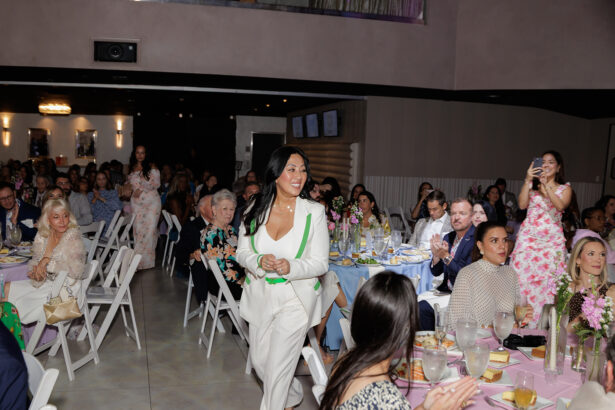 A woman in a white suit walks smiling through a crowded event hall with seated guests, many of whom are clapping or taking photos. Tables are decorated with flowers and food. The atmosphere is festive and celebratory. South Florida Business & Wealth