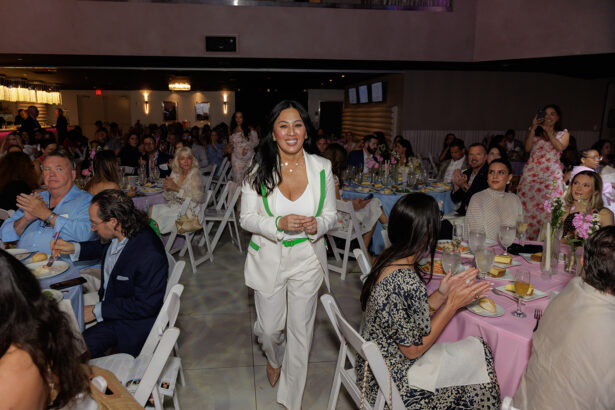 A woman in a white suit with green trim walks smiling between tables at a crowded indoor event while people seated around her clap and watch. The room is decorated with pastel colors and flowers. South Florida Business & Wealth
