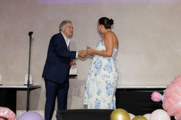 A man in a navy suit shakes hands with a woman in a strapless blue and white floral dress on a stage decorated with colorful balloons. The woman holds a plaque, and both are smiling. South Florida Business & Wealth