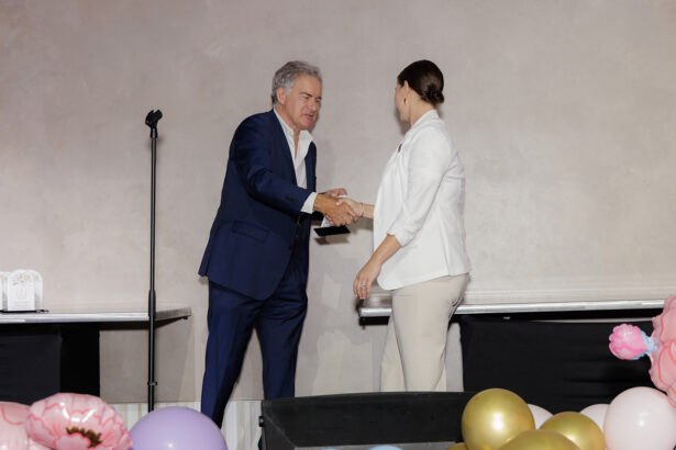 A man in a blue suit shakes hands with a woman in a white blazer on a stage decorated with colorful balloons, next to a microphone stand and two tables. South Florida Business & Wealth
