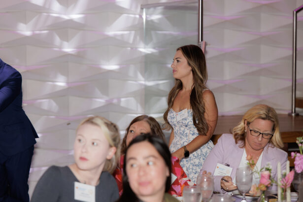 A group of women at an indoor event are seated at a table decorated with flowers. One woman stands nearby in a floral dress, while the others sit and look in various directions. The background features a textured, white wall. South Florida Business & Wealth
