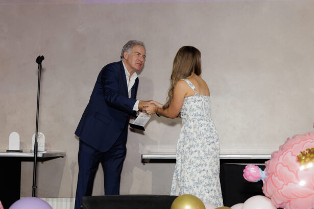 A man in a blue suit shakes hands with a woman in a white floral dress on a stage, with a microphone stand, pastel balloons, and pink flower decorations nearby. South Florida Business & Wealth