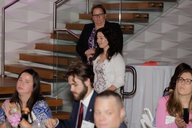 Two women stand near a staircase at an indoor event, while several people sit at tables in the foreground, some clapping. The setting appears formal, with name tags visible and floral decorations on the tables. South Florida Business & Wealth