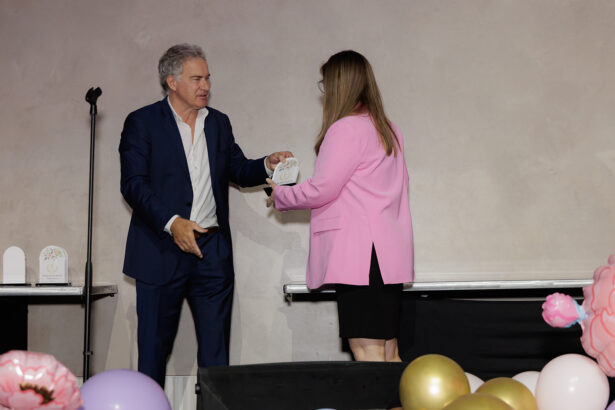 A man in a blue suit hands an award to a woman in a pink blazer on a stage decorated with colorful balloons, with a microphone stand visible in the background. South Florida Business & Wealth