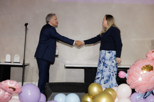 A man and woman in formal attire shake hands on stage, surrounded by colorful balloons and large decorative flowers. A microphone stand and award plaques are visible in the background. South Florida Business & Wealth