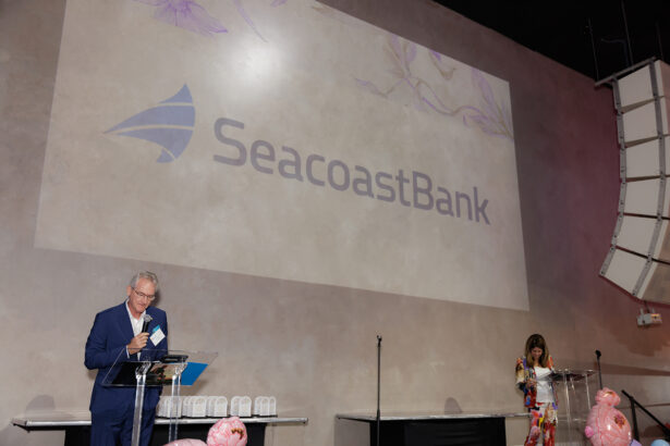 A man and a woman stand at separate podiums on stage with a large "Seacoast Bank" logo projected on the wall behind them. The podiums have transparent stands and pink decorations on the tables nearby. South Florida Business & Wealth