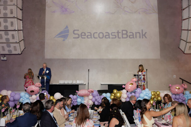 A formal event with guests seated at decorated tables, colorful balloons, and floral arrangements. Large SeacoastBank logo is projected on the wall behind two speakers at podiums on stage. South Florida Business & Wealth