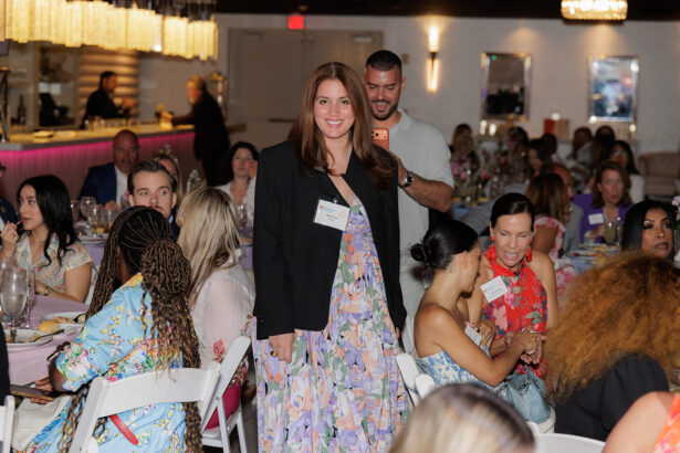 A woman in a floral dress and black blazer stands and smiles at a seated event in a decorated venue, surrounded by people at round tables with pastel tablecloths and elegant lighting. South Florida Business & Wealth