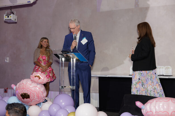 Three people stand on a stage decorated with pastel balloons. A man in a blue suit speaks at a clear podium while two women in colorful dresses look on and smile. There is a projector on the wall above them. South Florida Business & Wealth