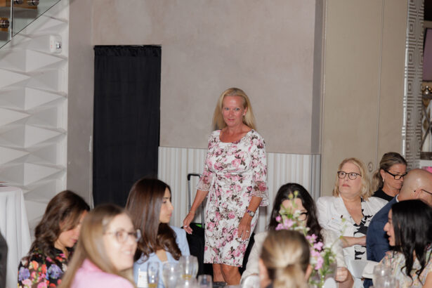 A woman in a floral dress stands and smiles in a room with seated guests, some looking toward her. The setting appears to be a formal event or gathering with tables and floral centerpieces. South Florida Business & Wealth