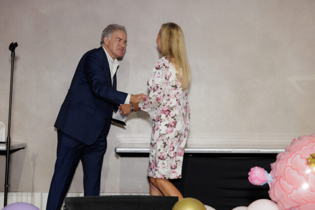 A man in a blue suit shakes hands with a woman in a floral dress on a stage. There is a microphone stand to the left and pastel-colored balloons to the right. Both are smiling and facing each other. South Florida Business & Wealth