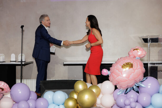 A man in a navy suit and a woman in a red dress shake hands on stage, surrounded by pastel and gold balloon decorations and large flower balloons. An acrylic podium and award plaques are visible in the background. South Florida Business & Wealth