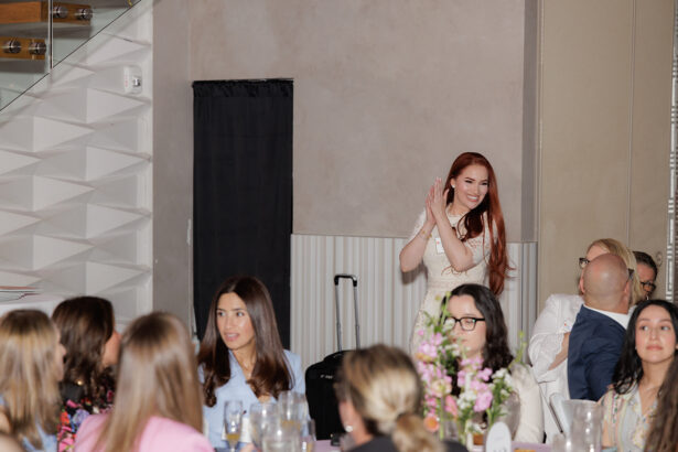 A woman with long red hair stands and claps, smiling, while a group of people seated at tables in the foreground look toward her in a brightly lit indoor setting. South Florida Business & Wealth