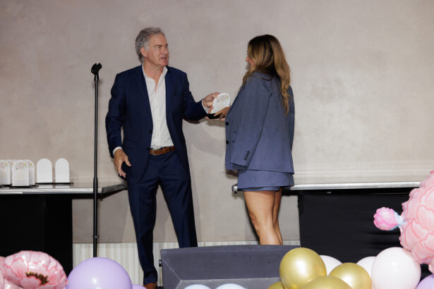 A man in a navy suit hands an award to a woman in a blue blazer and shorts on a stage decorated with pastel-colored balloons. A microphone stands nearby, and more awards rest on a table. South Florida Business & Wealth