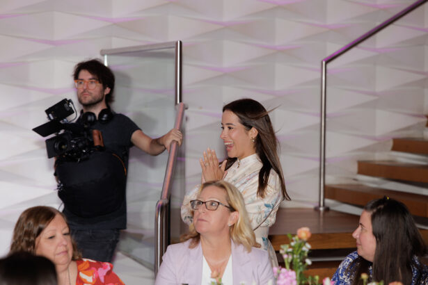 A smiling woman stands with her hands clasped near her face, looking excited. Seated women and a cameraman are nearby. The setting appears to be indoors with modern decor and a staircase in the background. South Florida Business & Wealth