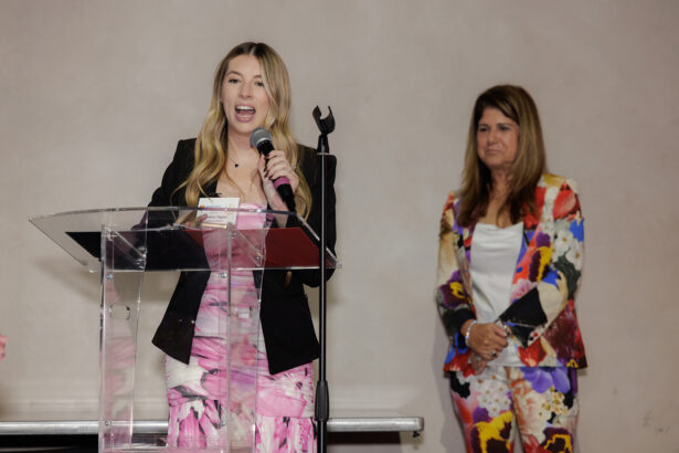 A woman in a black blazer and pink patterned dress speaks at a clear podium with a microphone, while another woman in a colorful floral suit stands in the background, watching. South Florida Business & Wealth