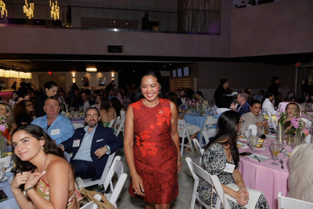 A woman in a red dress smiles as she walks between tables at a formal event. Guests seated at round tables with floral centerpieces watch her, and the room is filled with people dressed in formal attire. South Florida Business & Wealth