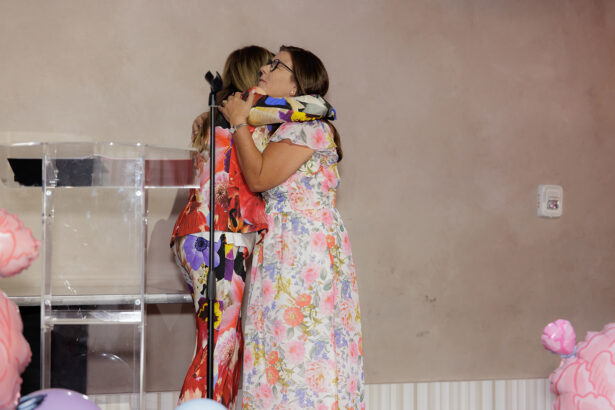 Two women in colorful floral dresses hug on a stage near a clear podium with a microphone. The background is plain, and pink decorations are visible at the bottom of the image. South Florida Business & Wealth