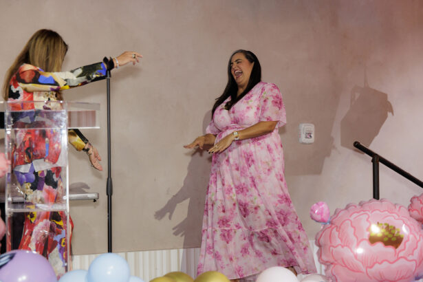 A woman in a pink floral dress walks onto a stage smiling, as another woman in a colorful dress reaches out to greet her. Balloons are visible on the floor near the stage. South Florida Business & Wealth