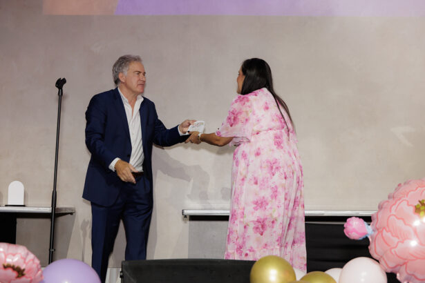 A man in a navy suit hands an award to a woman in a pink floral dress on stage, with decorative balloons in the foreground. South Florida Business & Wealth