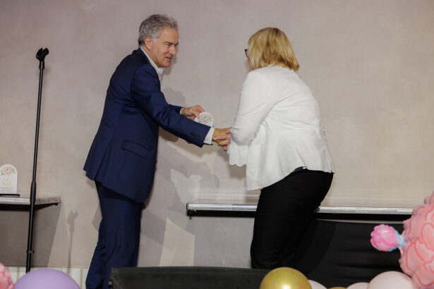 A man in a blue suit and a woman in a white blouse exchange an award on a stage decorated with balloons, with a microphone stand and table in the background. South Florida Business & Wealth