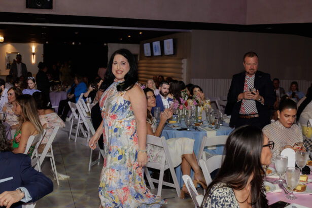 A woman in a floral dress smiles while walking between tables at a formal event. Guests seated around decorated tables are clapping, eating, and taking photos. The room is warmly lit, with several people focused on the woman. South Florida Business & Wealth