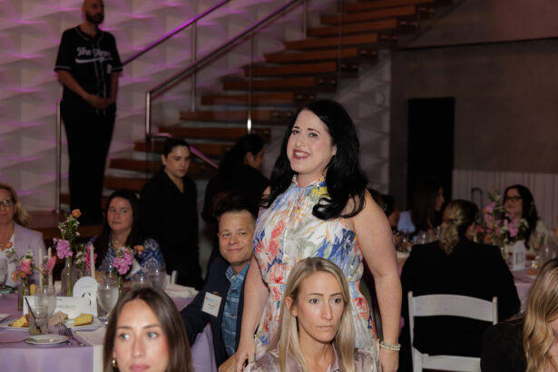 A woman in a floral dress stands and smiles in a room filled with seated people at round tables decorated with flowers; a staircase and people in the background. South Florida Business & Wealth