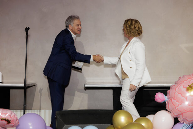 A man in a navy suit and a woman in a white suit shake hands on a stage decorated with colorful balloons and flowers, with a microphone stand in the background. South Florida Business & Wealth