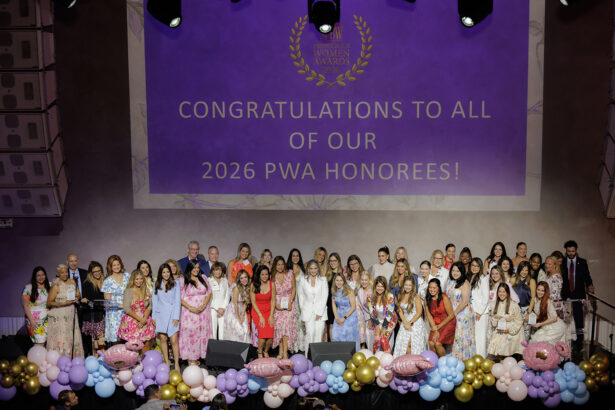 A large group of women and a few men pose on stage in front of a purple screen that reads, "Congratulations to all of our 2026 PWA Honorees!" Pink and purple balloons decorate the stage front. South Florida Business & Wealth