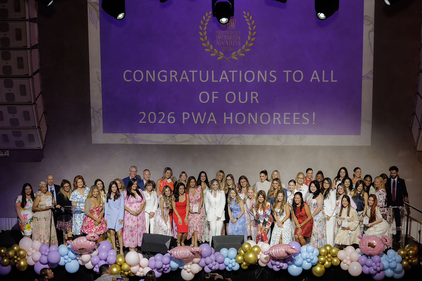 A large group of women and a few men stand on stage in front of a purple screen that reads, "Congratulations to all of our 2026 PWA Honorees!" The stage is decorated with colorful balloons and flowers. South Florida Business & Wealth