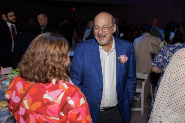 A man in a blue blazer with a pink flower boutonniere smiles and talks to a woman in a red floral top at a crowded indoor event. People mingle in the dimly lit background. South Florida Business & Wealth