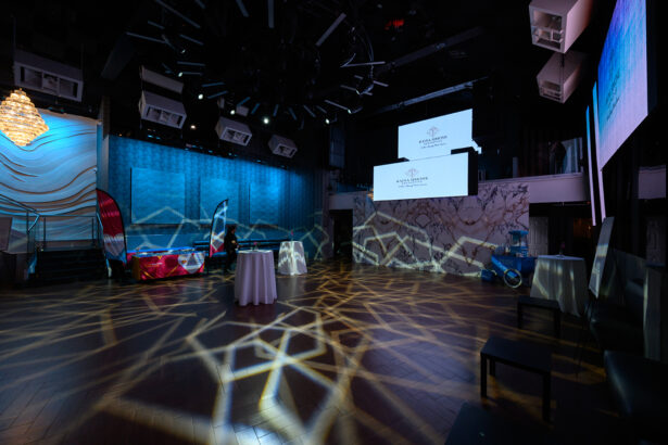 A dimly lit event space with patterned light projections on the wooden floor, a small round table with a white tablecloth at the center, banners and displays along the walls, and large projection screens above the stage area. South Florida Business & Wealth