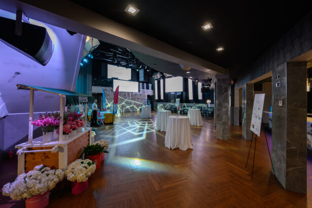 A modern event space with wooden floors, round cocktail tables with white tablecloths, wall-mounted screens, decorative lighting, and a flower cart filled with pink and white flowers. The area is ready for guests. South Florida Business & Wealth