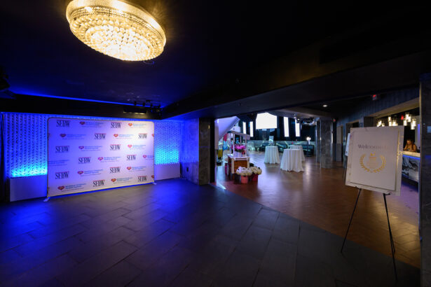 Spacious event venue with dim lighting, a chandelier, step-and-repeat banner on the left, welcome sign on an easel to the right, and tables with white tablecloths in the background. Blue and purple lights illuminate the room. South Florida Business & Wealth
