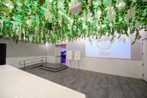 A modern event space with a gray tiled floor, white walls, and a ceiling covered in green leaves and hanging white flowers. Two poster stands and a projected award logo are visible near an entrance. South Florida Business & Wealth