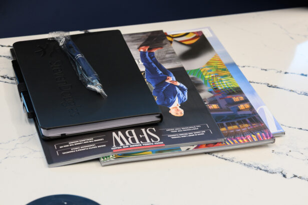 A black notebook with a pen on top rests on a marble surface, next to a stack of colorful magazines, one featuring a well-dressed person and vibrant outdoor scenes on its cover. South Florida Business & Wealth