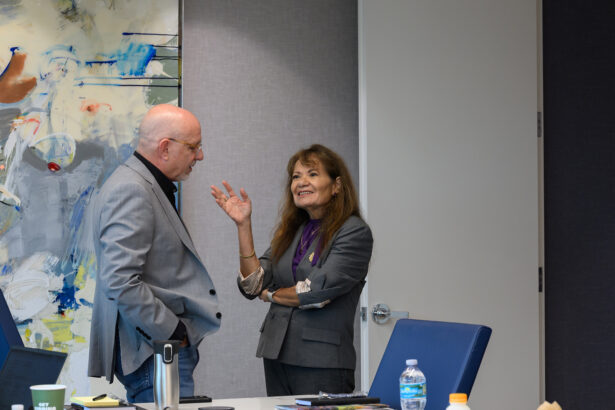 Two people in business attire stand and talk in a modern office near a table with drinks. One person, gesturing with a hand, smiles while the other listens attentively. Abstract art hangs on the wall in the background. South Florida Business & Wealth